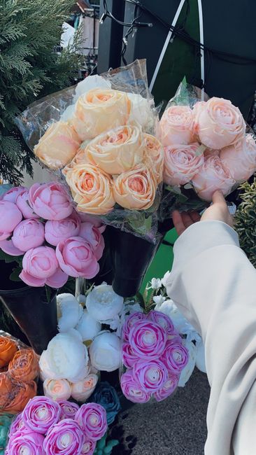 A person wearing a white long-sleeve garment holds a bouquet of peach and pink roses wrapped in clear plastic, showcasing their delicate petals and fresh green stems. Surrounding the bouquet are sever
