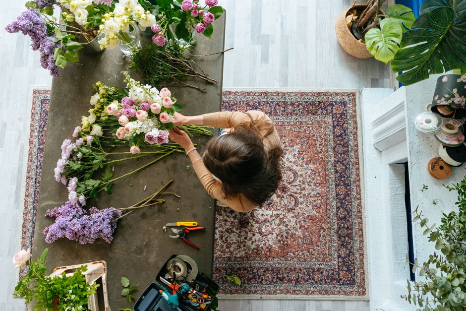 An overhead view of a woman arranging a floral bouquet at a grey work table in a well-lit room with a traditional red and blue patterned rug. She is selecting pink and white roses, peonies, and purple