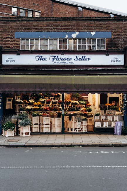 The exterior of The Flower Seller shop on Muswell Hill, featuring a white sign with blue cursive lettering and contact details, situated beneath a weathered blue awning on a brick building. The displa