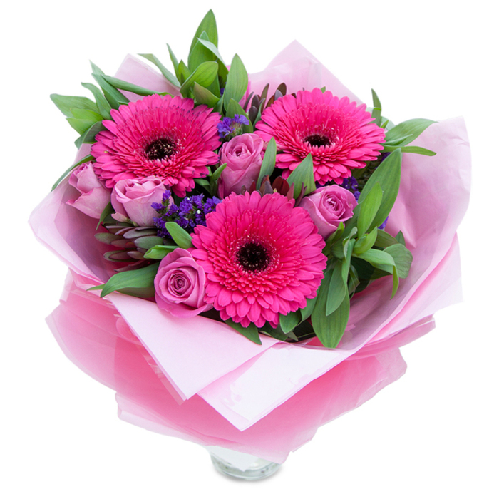 I see this arrangement as an elegant study in pinks and texture: three vivid fuchsia Gerbera daisies anchor the composition with their large, symmetrical heads and dark centres, while a cluster of lavender-pink roses nestle in close, their soft, tightly furled petals offering a gentle, romantic counterpoint. Sprigs of purple statice punctuate the design, introducing a crisp, bristly texture and a cooler violet hue that enhances the warmth of the pinks. The underlying greenery - broad leaves that resemble salal or eucalyptus - provides a deep green backdrop, its subtle sheen and fresh scent rooting the bouquet in a natural, garden-derived freshness. The flowers are wrapped in layers that read as both protective and ornamental: an inner, soft matte pale pink tissue cushions the stems, and an outer, slightly crinkled translucent cellophane in a matching fuchsia-pink adds a touch of shimmer and polish. The top rim of a clear glass vase is visible beneath the wrapping, suggesting the bouquet is intended for immediate display in a clear vessel. Under crisp daylight the colours appear saturated and joyful, and the interplay of textures - silky petals, papery statice, glossy foliage - invites touch as much as it does sight. As a local florist, I picture this arrangement brightening a mantelpiece in Collier Row village, greeting visitors in a Romford porch, or arriving as a thoughtful anniversary or birthday gesture for someone who appreciates bold colour with refined detail.