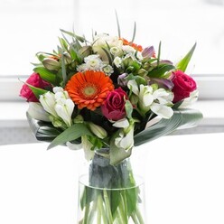A visual feast of colour and texture, this exquisite floral arrangement embodies the artistry of Collier Row's local floristry. Creamy peach gerberas, encircling their deep brown hearts, are nestled beside radiant orange cousins that blaze like the last light of day. Interspersed throughout are sumptuous magenta roses-each petal unfurling with layered richness-while clusters of white alstroemeria and tiny spray roses add splashes of brightness and subtle aroma. Sprigs of pink and delicate white blossoms lend movement and whimsy, evoking the sense of a well-tended garden bordering Mawney Road in early spring. Broad, glossy aspidistra leaves are gently curved and wrapped around the bouquet's base, forming a verdant, sculptural collar that cradles the vibrant blooms within. Smaller pointed leaves and airy greenery dart between each flower, introducing lively textures and contrast. Bathed in gentle, diffused light reminiscent of a Collier Row morning, every colour seems heightened and every petal inviting-infusing the bouquet with fresh energy and positivity. Resting in a tall, slender clear glass vase, the entire arrangement radiates delicate balance and an understated sophistication. This bouquet would be perfectly at home as a centrepiece for celebrations, anniversaries, or as a thoughtful gesture for new beginnings-a tangible symbol of care and optimism drawn from the heart of Collier Row's floral community.